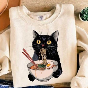 May include: A cream-colored sweatshirt with a black cat eating ramen noodles from a pink bowl. The cat has yellow eyes and a black nose.