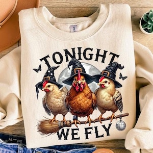 May include: Cream-colored sweatshirt with a graphic of three chickens wearing witch hats, perched on a broomstick, with the text "TONIGHT WE FLY." The design includes a full moon and bats. The sweatshirt is paired with blue denim shorts and a brown hat.