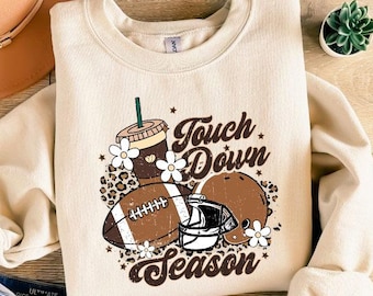 May include: Cream-colored sweatshirt with a football-themed graphic. The design includes a football, helmet, coffee cup, and the text "Touch Down Season." The sweatshirt is paired with blue denim shorts, a brown hat, and sandals.