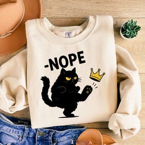 May include: Cream-coloured sweatshirt featuring the word "NOPE" above a black cat illustration wearing a gold crown. The cat is kicking its leg up. A brown hat and blue jeans are also in the image.