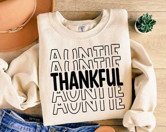 Può includere: Una felpa color crema con la scritta "Auntie Thankful" stampata in nero. Il testo è ripetuto più volte in uno schema impilato.
