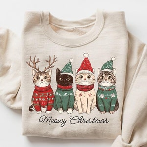 May include: A beige sweatshirt with a Christmas-themed design featuring four cats wearing Santa hats and sweaters. The text "Meowy Christmas" is printed below the cats.