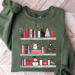 May include: A green sweatshirt with a Christmas-themed design featuring a bookshelf with books, candles, a snowman, Santa Claus, a cat, and a Christmas tree.