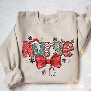 May include: A beige sweatshirt with a Christmas-themed design that says "Nurse" in colorful letters with a red bow and a stethoscope.