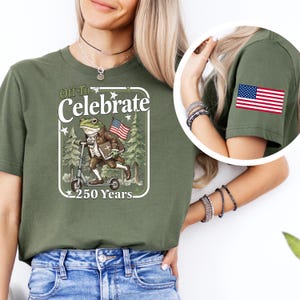 May include: Olive green t-shirt featuring a graphic of a frog riding a scooter, holding an American flag, and the text "Off To Celebrate 250 Years." An American flag patch is on the sleeve.