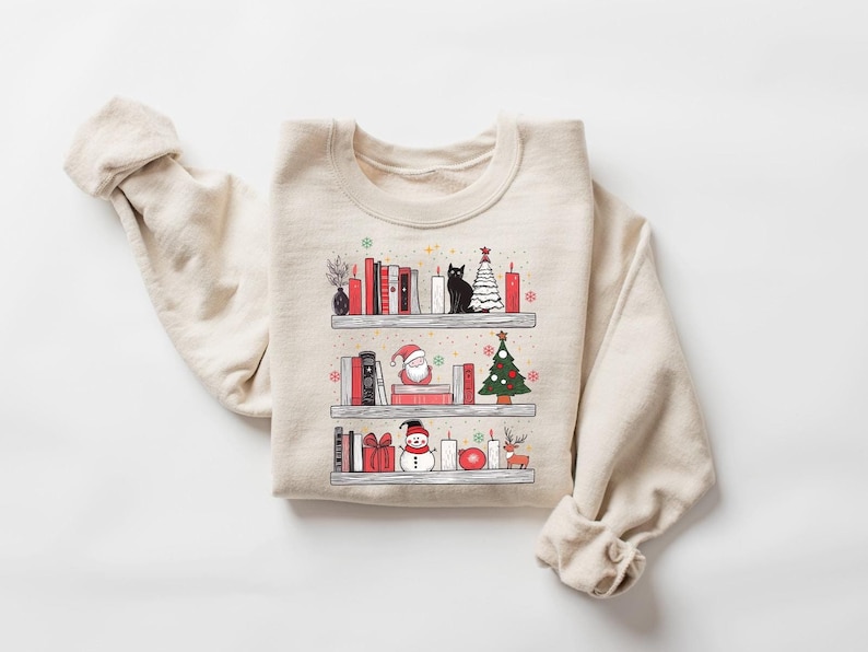 May include: A beige sweatshirt with a Christmas-themed design featuring a bookshelf with books, a cat, a Christmas tree, a snowman, a Santa Claus, and candles.