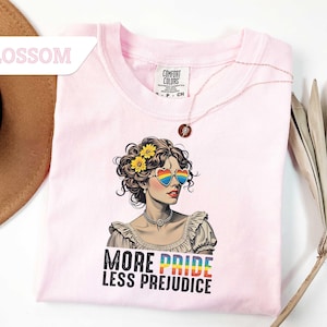 May include: A light pink t-shirt featuring a graphic of a woman wearing heart-shaped rainbow sunglasses and flowers in her hair. The text "MORE PRIDE LESS PREJUDICE" is printed below the graphic. A gold necklace is draped across the shirt.