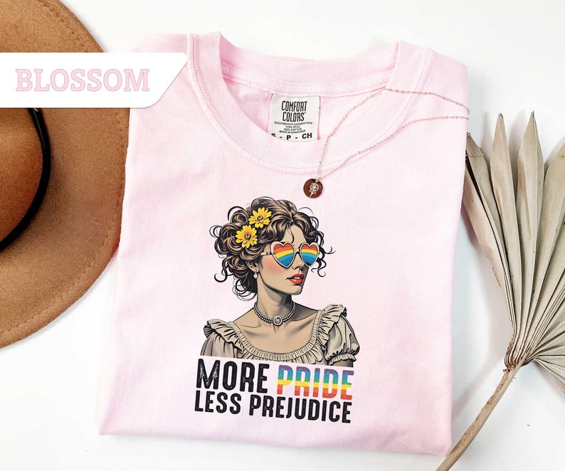 May include: A light pink t-shirt featuring a graphic of a woman wearing heart-shaped rainbow sunglasses and flowers in her hair. The text "MORE PRIDE LESS PREJUDICE" is printed below the graphic. A gold necklace is draped across the shirt.