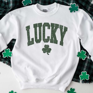 Peut inclure: Sweat-shirt blanc avec le mot "LUCKY" imprim&eacute; en vert sur le devant. Le mot est dans une police de caract&egrave;res de style vintage. Un tr&egrave;fle vert se trouve sous le mot.