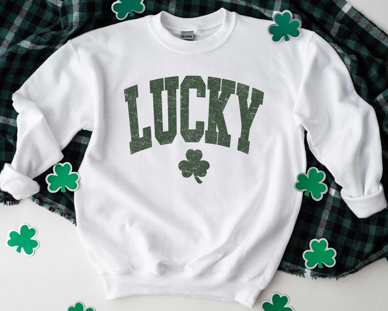 Peut inclure: Sweat-shirt blanc avec le mot "LUCKY" imprim&eacute; en vert sur le devant. Le mot est dans une police de caract&egrave;res de style vintage. Un tr&egrave;fle vert se trouve sous le mot.