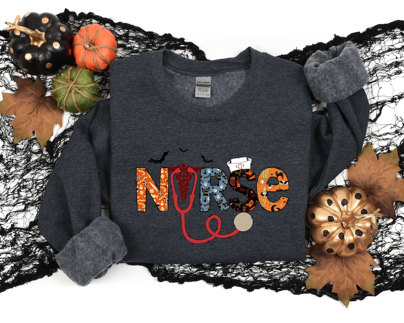 Pu&ograve; includere: Una felpa grigia scura con una grafica "INFERMIERA" a tema Halloween. Le lettere sono composte da varie icone di Halloween, tra cui pipistrelli, fantasmi e zucche. Un stetoscopio rosso &egrave; incluso nel design.