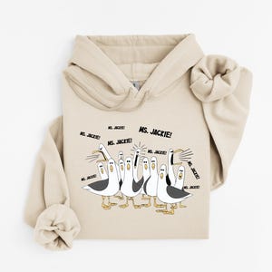 May include: A beige hoodie featuring a cartoon illustration of seagulls with the text "Ms. Jackie!". The seagulls are depicted in black, white, and yellow, with the text in black. The hoodie has a drawstring hood and a kangaroo pocket.