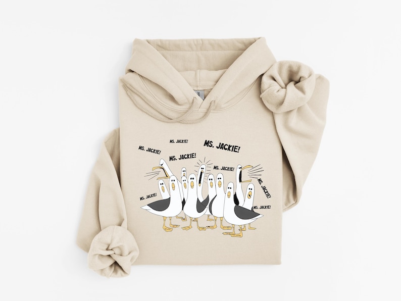 May include: A beige hoodie featuring a cartoon illustration of seagulls with the text "Ms. Jackie!". The seagulls are depicted in black, white, and yellow, with the text in black. The hoodie has a drawstring hood and a kangaroo pocket.