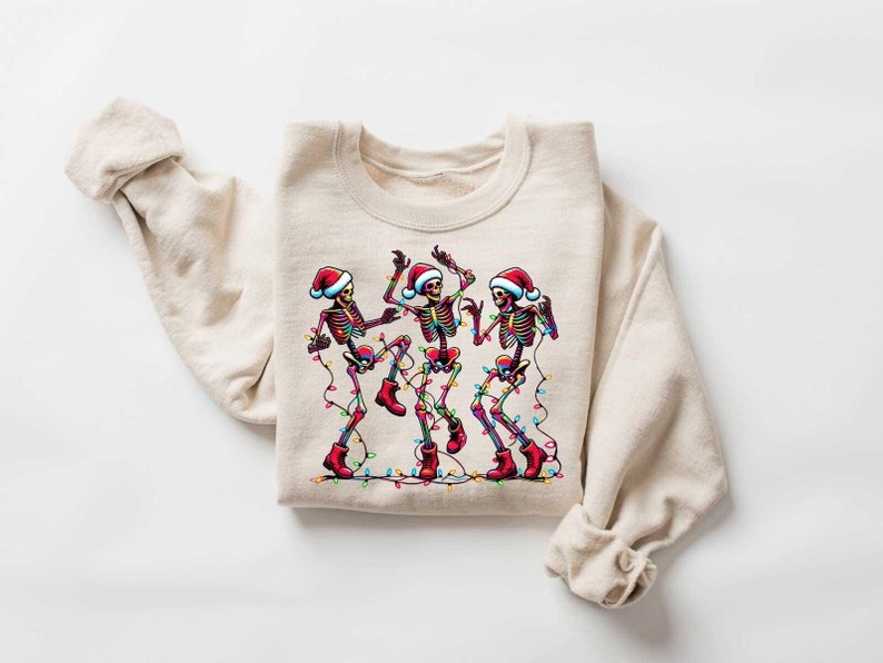 May include: A beige sweatshirt with a festive design of three skeletons wearing Santa hats and dancing with Christmas lights.