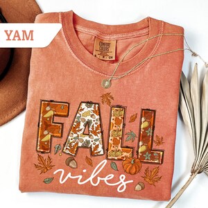 May include: A coral-colored t-shirt with the words "FALL vibes" in a decorative font. The word "FALL" is filled with fall-themed illustrations of leaves, acorns, and pumpkins. A gold necklace and a brown hat are also visible.
