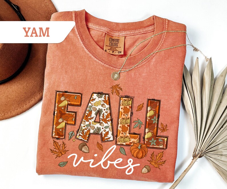 May include: A coral-colored t-shirt with the words "FALL vibes" in a decorative font. The word "FALL" is filled with fall-themed illustrations of leaves, acorns, and pumpkins. A gold necklace and a brown hat are also visible.