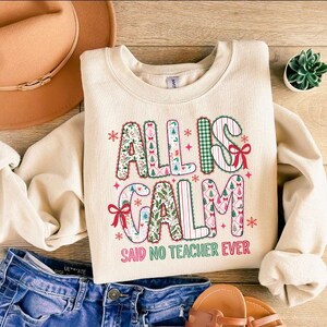 May include: Cream-colored sweatshirt with the text "ALL IS CALM SAID NO TEACHER EVER" in a festive, patterned design. The sweatshirt is paired with blue jeans, a brown hat, and sandals, suggesting a casual, everyday style.