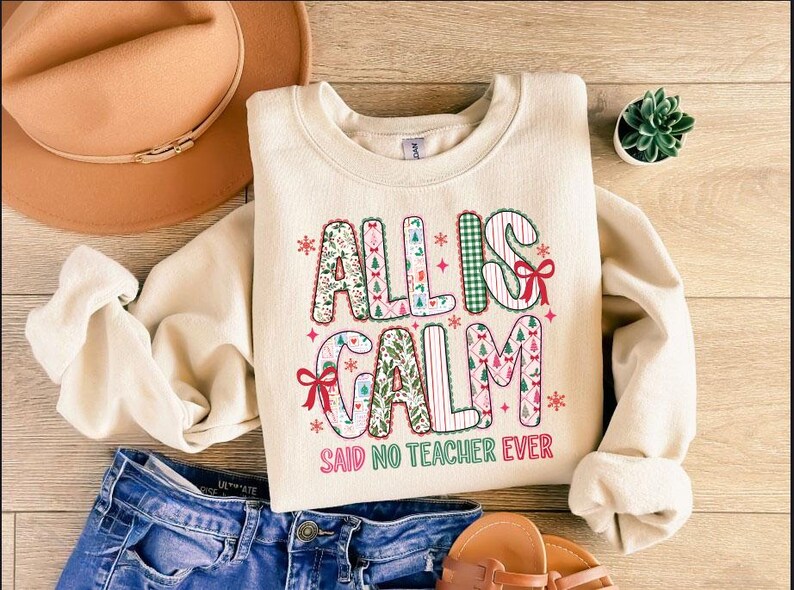 May include: Cream-colored sweatshirt with the text "ALL IS CALM SAID NO TEACHER EVER" in a festive, patterned design. The sweatshirt is paired with blue jeans, a brown hat, and sandals, suggesting a casual, everyday style.