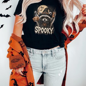 May include: A black t-shirt featuring a graphic of a raccoon wearing a witch hat, holding a pumpkin and a drink, with a full moon background and the text SPOOKY. The shirt is paired with light blue distressed denim jeans and an orange cardigan.
