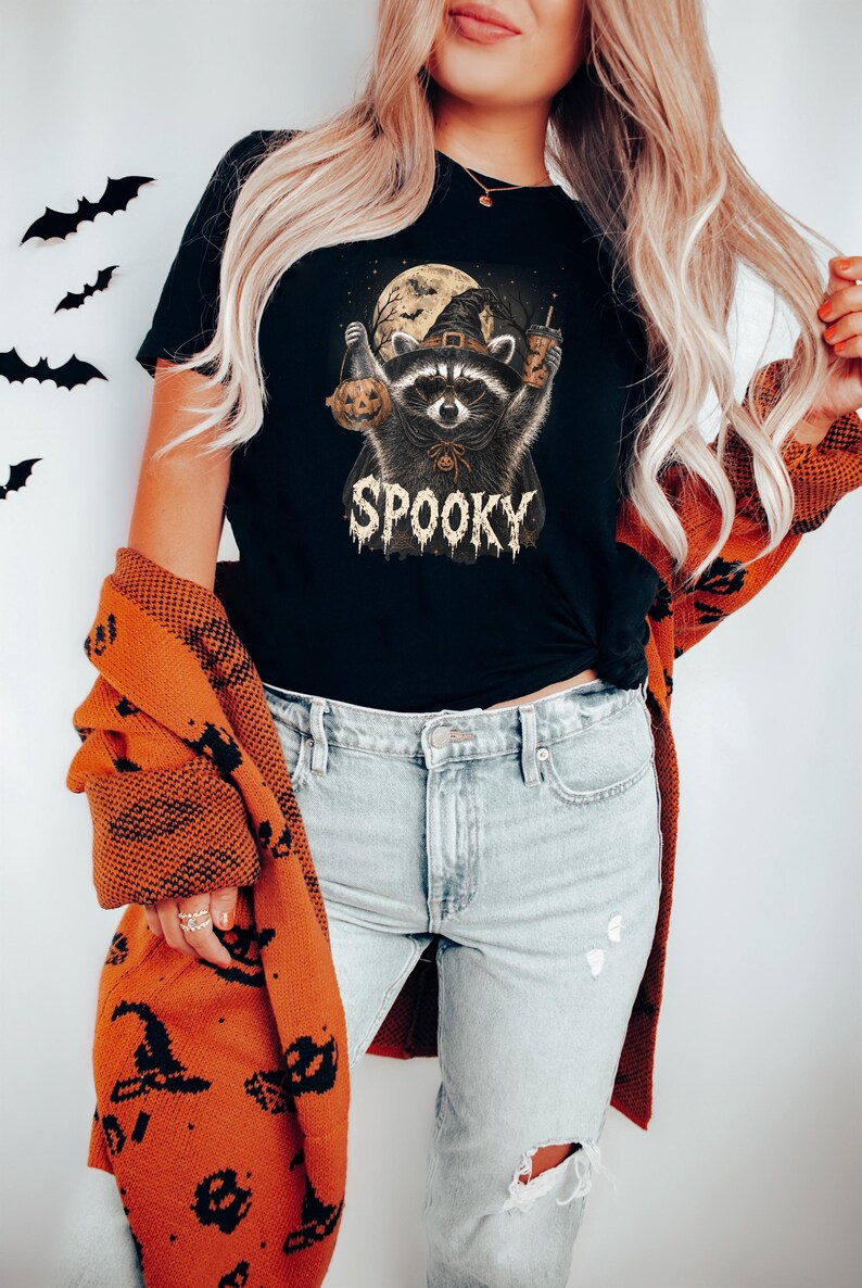 May include: A black t-shirt featuring a graphic of a raccoon wearing a witch hat, holding a pumpkin and a drink, with a full moon background and the text SPOOKY. The shirt is paired with light blue distressed denim jeans and an orange cardigan.