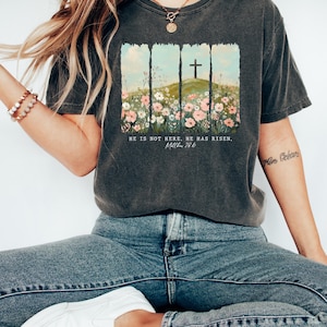 May include: A dark gray t-shirt with a graphic of a cross on a hill with flowers. The text "He is not here. He has risen. Matthew 28:6" is printed below the graphic.