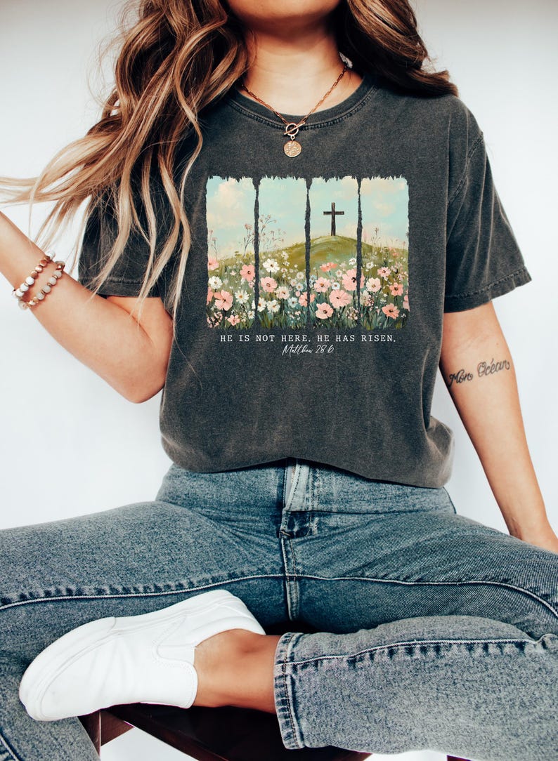 May include: A dark gray t-shirt with a graphic of a cross on a hill with flowers. The text "He is not here. He has risen. Matthew 28:6" is printed below the graphic.