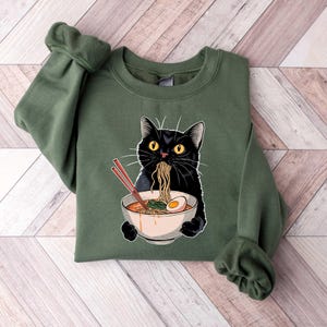 May include: A green sweatshirt with a black cat eating ramen noodles from a bowl. The cat has yellow eyes and is wearing a red collar.