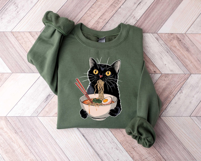 May include: A green sweatshirt with a black cat eating ramen noodles from a bowl. The cat has yellow eyes and is wearing a red collar.