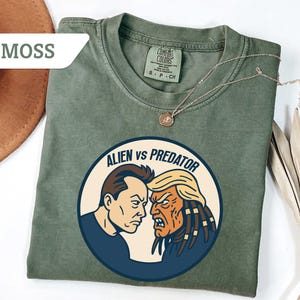May include: Moss green t-shirt with a round graphic featuring two opposing figures labeled "Alien vs Predator." The shirt has a crew neck and short sleeves. A gold necklace with a pendant is draped on the shirt. The shirt is made of a soft, comfortable material.