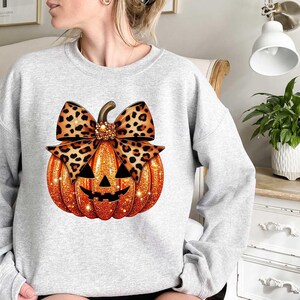 Peut inclure: Un sweat-shirt gris avec une citrouille orange scintillante orn&eacute;e d'un n&oelig;ud &agrave; imprim&eacute; l&eacute;opard. La citrouille a un visage de citrouille d'Halloween.