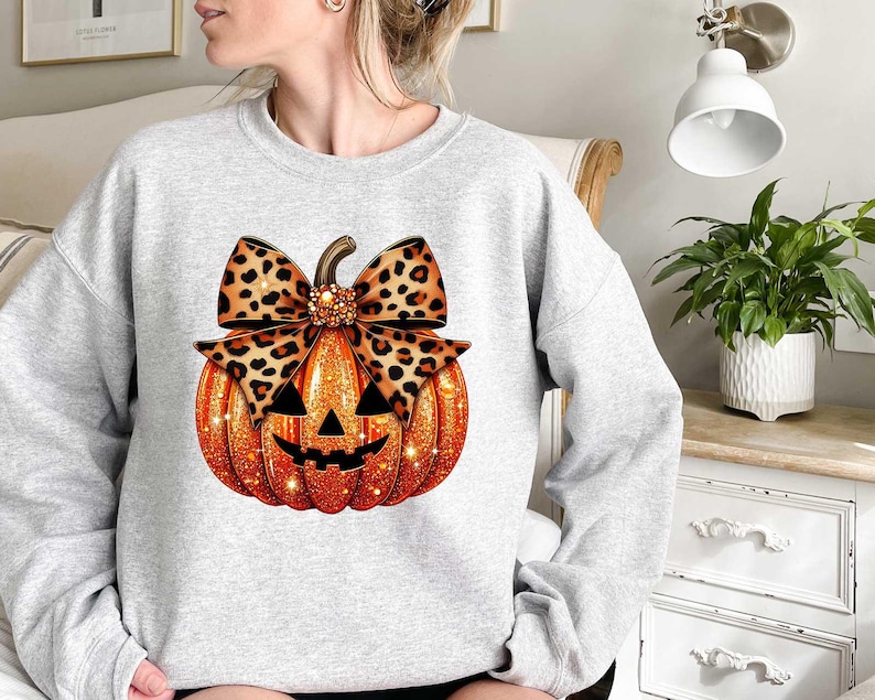 Peut inclure: Un sweat-shirt gris avec une citrouille orange scintillante orn&eacute;e d'un n&oelig;ud &agrave; imprim&eacute; l&eacute;opard. La citrouille a un visage de citrouille d'Halloween.