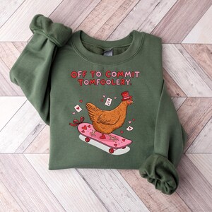 K&ouml;nnte beinhalten: Ein gr&uuml;nes Sweatshirt mit dem Text "OFF TO COMMIT TOMFOOLERY". Ein Cartoon-Huhn mit Hut f&auml;hrt auf einem rosa Skateboard mit Herzen. Das Design enth&auml;lt kleine Herzen und Umschl&auml;ge.