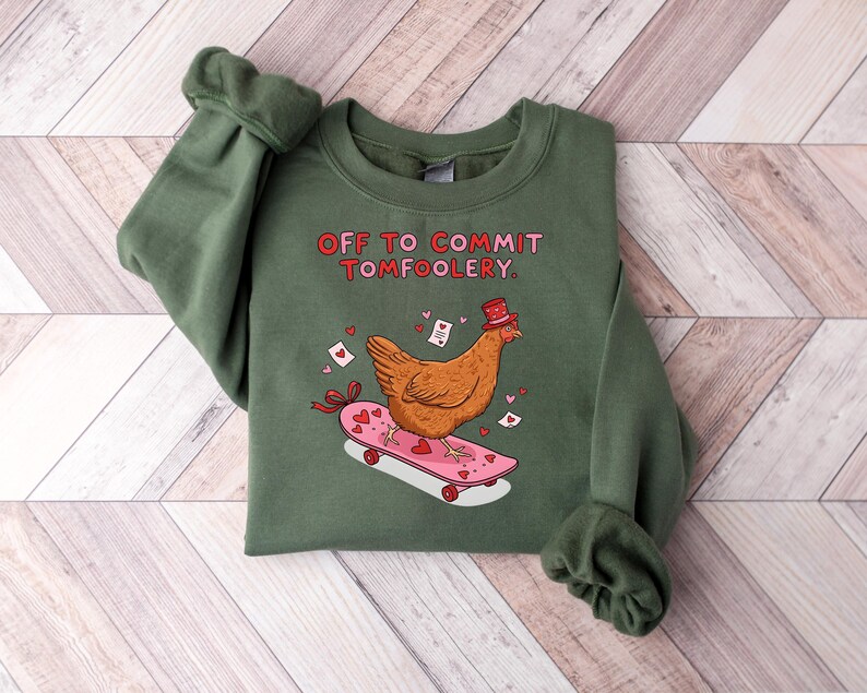K&ouml;nnte beinhalten: Ein gr&uuml;nes Sweatshirt mit dem Text "OFF TO COMMIT TOMFOOLERY". Ein Cartoon-Huhn mit Hut f&auml;hrt auf einem rosa Skateboard mit Herzen. Das Design enth&auml;lt kleine Herzen und Umschl&auml;ge.