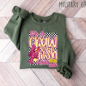 Puede incluir: Una sudadera verde con un gr&aacute;fico de un cangrejo de r&iacute;o rosa y el texto "It's a crawfish kinda day" en letras amarillas y rosas.