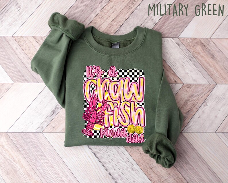 Puede incluir: Una sudadera verde con un gr&aacute;fico de un cangrejo de r&iacute;o rosa y el texto "It's a crawfish kinda day" en letras amarillas y rosas.