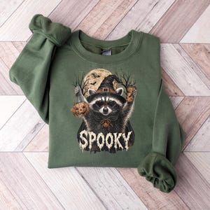 May include: An olive green crewneck sweatshirt featuring a graphic of a raccoon wearing a witch hat and cape. The raccoon holds a jack-o'-lantern and a beverage cup. Behind it is a full moon and bare trees. The word SPOOKY is printed below.