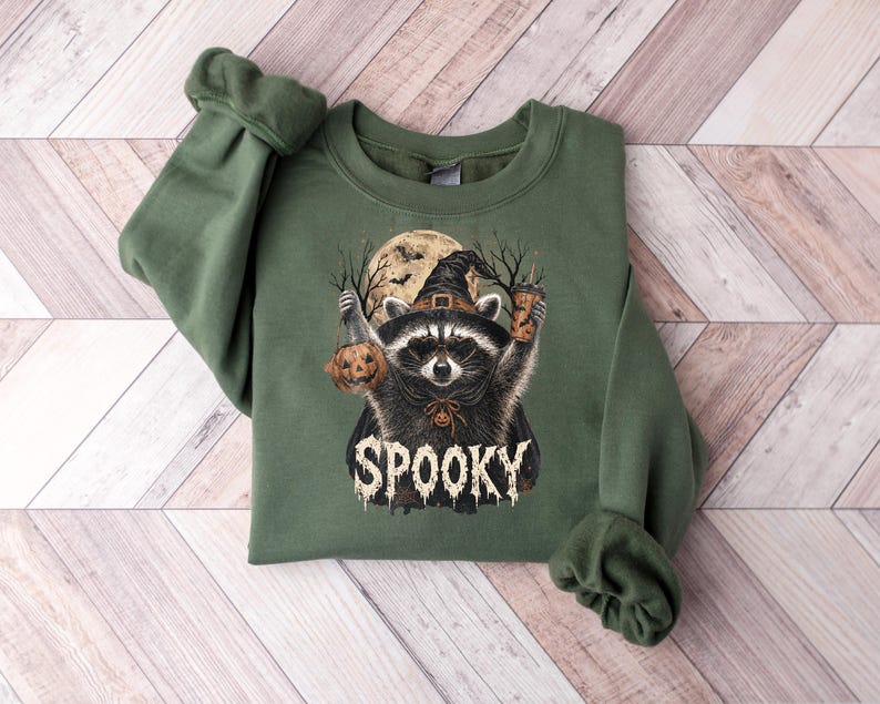 May include: An olive green crewneck sweatshirt featuring a graphic of a raccoon wearing a witch hat and cape. The raccoon holds a jack-o'-lantern and a beverage cup. Behind it is a full moon and bare trees. The word SPOOKY is printed below.