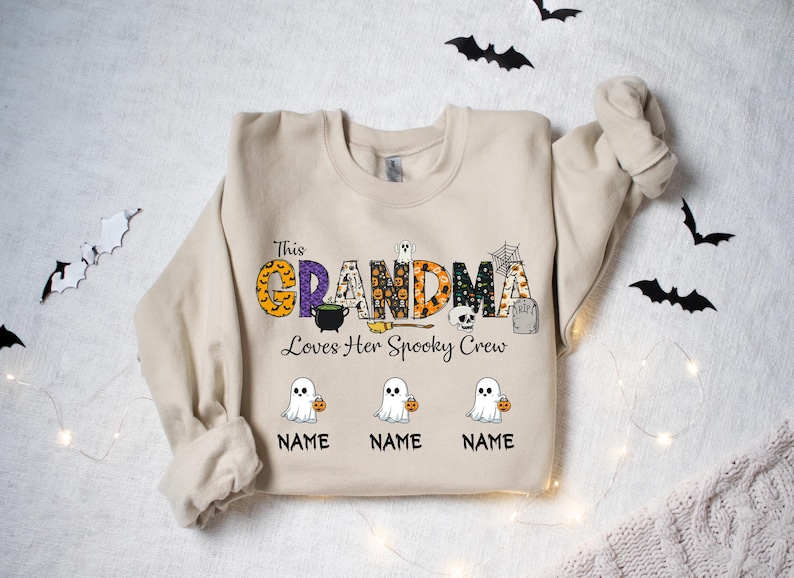 May include: Beige Halloween sweatshirt with the text "This Grandma Loves Her Spooky Crew." The word "GRANDMA" is in orange, purple, and black patterns. Below are three cartoon ghosts holding pumpkins, with space for names.