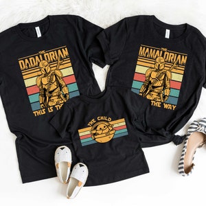 May include: Three black t-shirts with a retro design featuring a Mandalorian character. The shirts have the text "The Dadalorian This Is The Way", "The Mamalorian The Way", and "The Child".