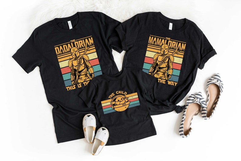 May include: Three black t-shirts with a retro design featuring a Mandalorian character. The shirts have the text "The Dadalorian This Is The Way", "The Mamalorian The Way", and "The Child".