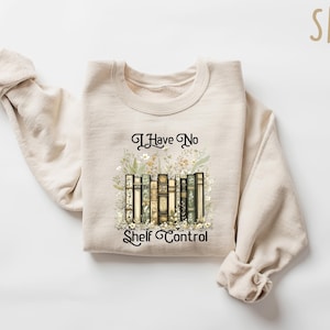 May include: A sand-colored sweatshirt with the text "I Have No Shelf Control" in black script. The graphic features a row of books with floral accents. The sweatshirt is made of a soft material.