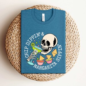 Pu&ograve; includere: Una maglietta blu con una grafica bianca di uno scheletro che beve una margarita e il testo "Chip Dippin' & Sippin' Margarita".