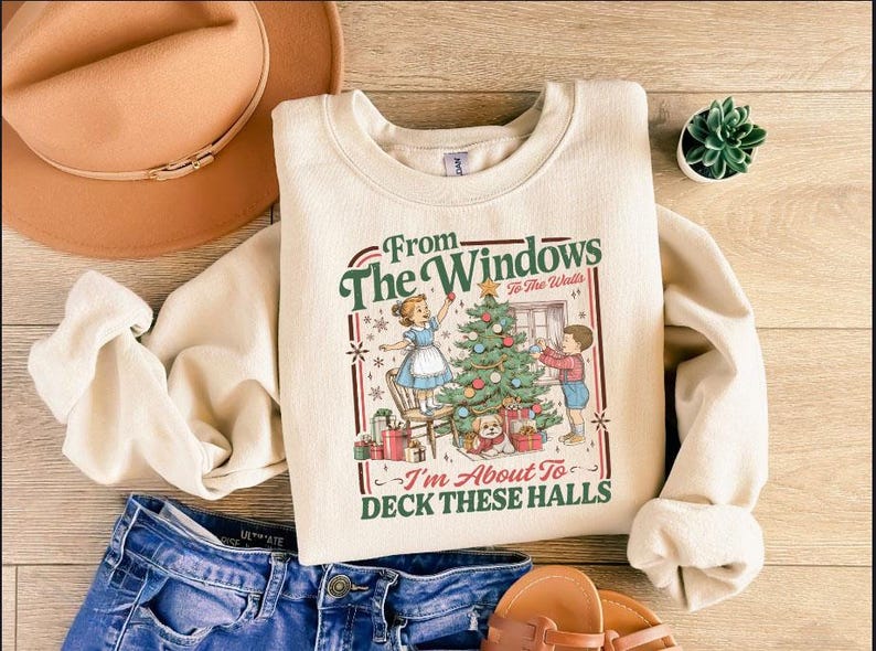 May include: Cream-colored sweatshirt with a Christmas-themed graphic. The graphic features children decorating a Christmas tree, with text that reads "From The Windows To The Walls, I'm About To Deck These Halls."