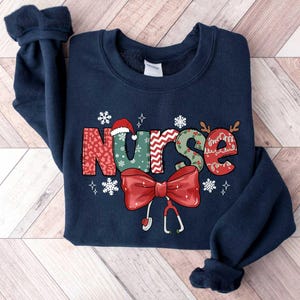 May include: Navy blue sweatshirt with a Christmas-themed design featuring the word "NURSE" in colorful, patterned letters with a red bow and a stethoscope.