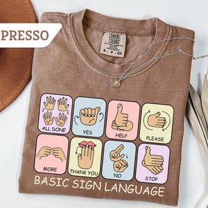 May include: Espresso-colored t-shirt with a graphic of basic sign language symbols. The shirt features the words "ALL DONE", "YES", "HELP", "PLEASE", "MORE", "THANK YOU", "NO", and "STOP". A silver necklace with a pendant is draped across the shirt.