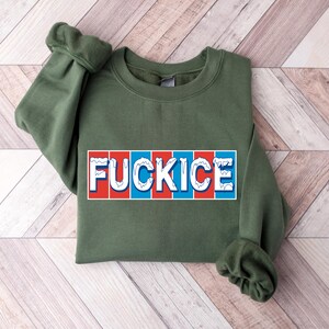 以下が含まれることがあります： 胸に「FUCKICE」の文字が長方形のデザインでプリントされたオリーブグリーンのスウェットシャツ。テキストは白で、赤と青の縦縞模様。袖口は折り返されています。