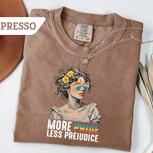 May include: Espresso-colored t-shirt featuring a graphic of a woman with heart-shaped rainbow sunglasses and yellow flowers in her hair. The text "MORE PRIDE LESS PREJUDICE" is printed below the graphic. A silver necklace is draped across the shirt.
