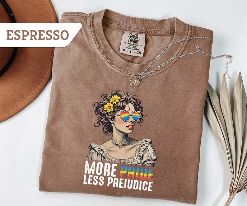 May include: Espresso-colored t-shirt featuring a graphic of a woman with heart-shaped rainbow sunglasses and yellow flowers in her hair. The text "MORE PRIDE LESS PREJUDICE" is printed below the graphic. A silver necklace is draped across the shirt.