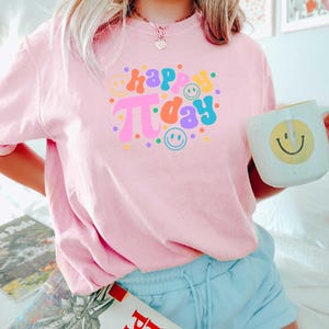 May include: A pink t-shirt featuring the text "happy pi day" in colourful, retro-style lettering. The shirt is decorated with colourful dots and smiley faces.