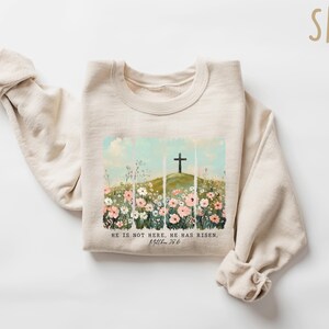 May include: A beige sweatshirt with a graphic print of a cross on a hill with flowers. The text "He is not here. He has risen. Matthew 28:6" is printed below the graphic.